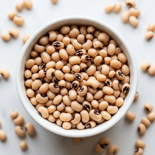 A white bowl filled with a single layer of cooked black-eyed peas, showing their pale beige color with distinctive black spots and a soft, slightly shiny texture. The bowl sits on a white marbled surface scattered with raw black-eyed peas around it, adding a contrast with their matte finish. The focus is on the bowl's content, capturing the small round legumes closely with natural light softening the scene. photo taken with an iphone --ar 4:5 --v 7