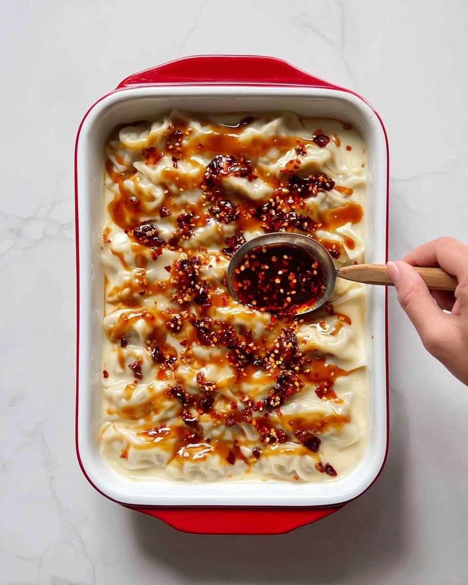 The image shows a white rectangular dish with a red rim filled with a creamy white layer of soft, folded dumplings covering the bottom. On top, there are uneven drizzles of light brown sauce and bright red chili oil with seeds and bits of chili scattered unevenly over the dumplings. A woman's hand holding a spoon is gently spreading the chili oil over the top right corner of the dish. The dish sits on a white marbled surface. photo taken with an iphone --ar 4:5 --v 7