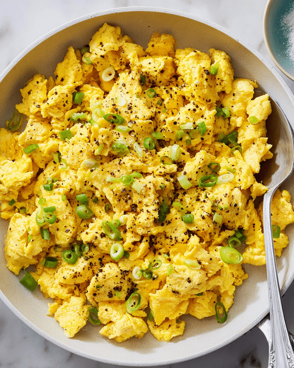 A close-up view of a white pan filled with fluffy scrambled eggs that are bright yellow with a slightly uneven texture, showing soft curds and small chunks. The eggs are sprinkled with small pieces of fresh, sliced green onions and a light dusting of black pepper, adding little pops of green and black on top. The pan sits on a white marbled surface with part of a silver spoon handle visible on the right side. Photo taken with an iphone --ar 4:5 --v 7
