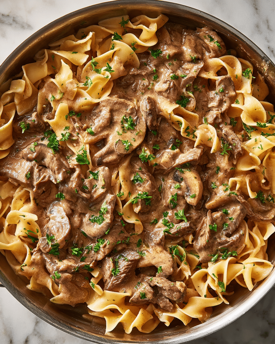 This dish shows a close-up of beef stroganoff in a large metal pan. The base layer has wide, flat egg noodles with a light golden color and soft, slightly curled edges. On top, there are tender slices of brown beef mixed with thinly sliced mushrooms coated in a creamy, brown sauce. Small green parsley pieces are sprinkled evenly across the surface, adding a fresh contrast. The sauce has a rich and smooth texture, covering the noodles and beef uniformly. The image captures the dish in warm lighting, highlighting the creaminess and the textures of the meat and noodles, on a background with a white marbled texture. Photo taken with an iphone --ar 4:5 --v 7