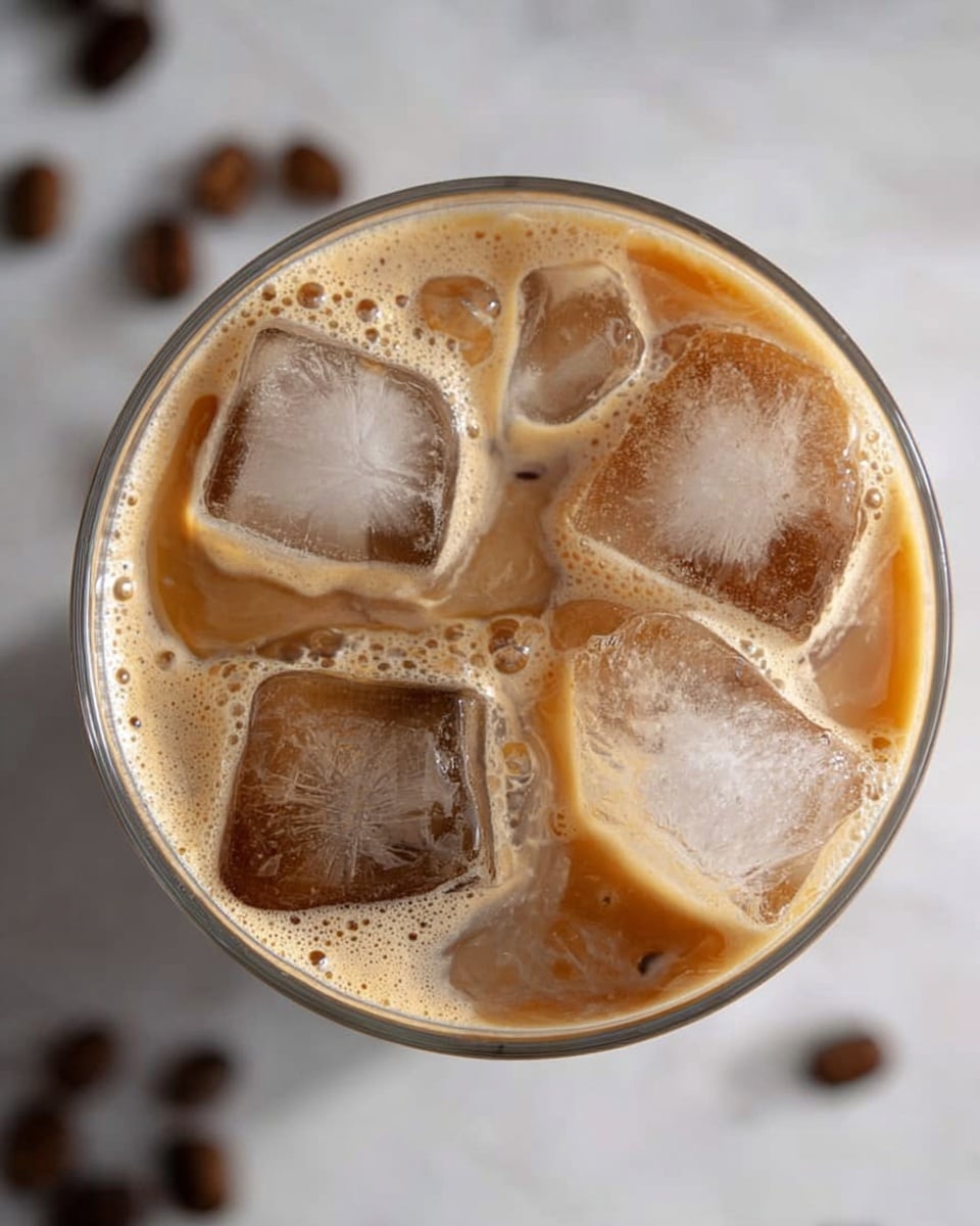 A clear glass filled with iced coffee viewed from above, showing four large irregularly shaped ice cubes in light brown coffee with creamy froth floating around and bubbles scattered on the surface, all placed on a white marbled texture with a few blurred coffee beans nearby, photo taken with an iphone --ar 4:5 --v 7