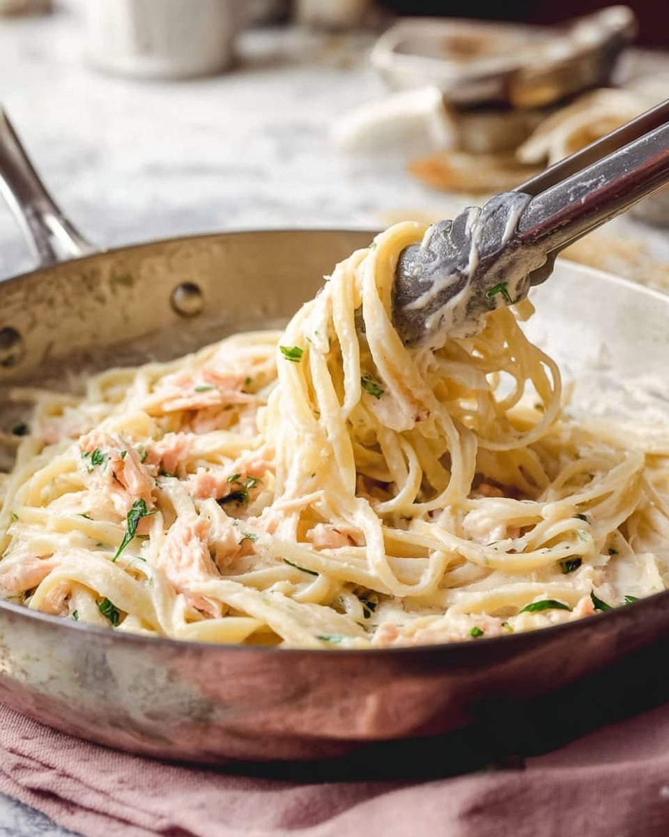 A white bowl with a blue rim filled with creamy pasta, featuring thick, pale yellow noodles mixed with pale pink chunks of salmon, topped with finely chopped green herbs and a light sprinkling of grated cheese. Behind this bowl, another similar bowl of the same pasta is partially visible with a fork resting on its edge. In the background, a white saucepan contains more pasta, and two small golden-yellow blocks of cheese sit on a white napkin next to a metal grater. A soft pink cloth is laid beside the front bowl, resting on a surface with a white marbled texture. A silver fork lies nearby in the foreground. photo taken with an iphone --ar 4:5 --v 7