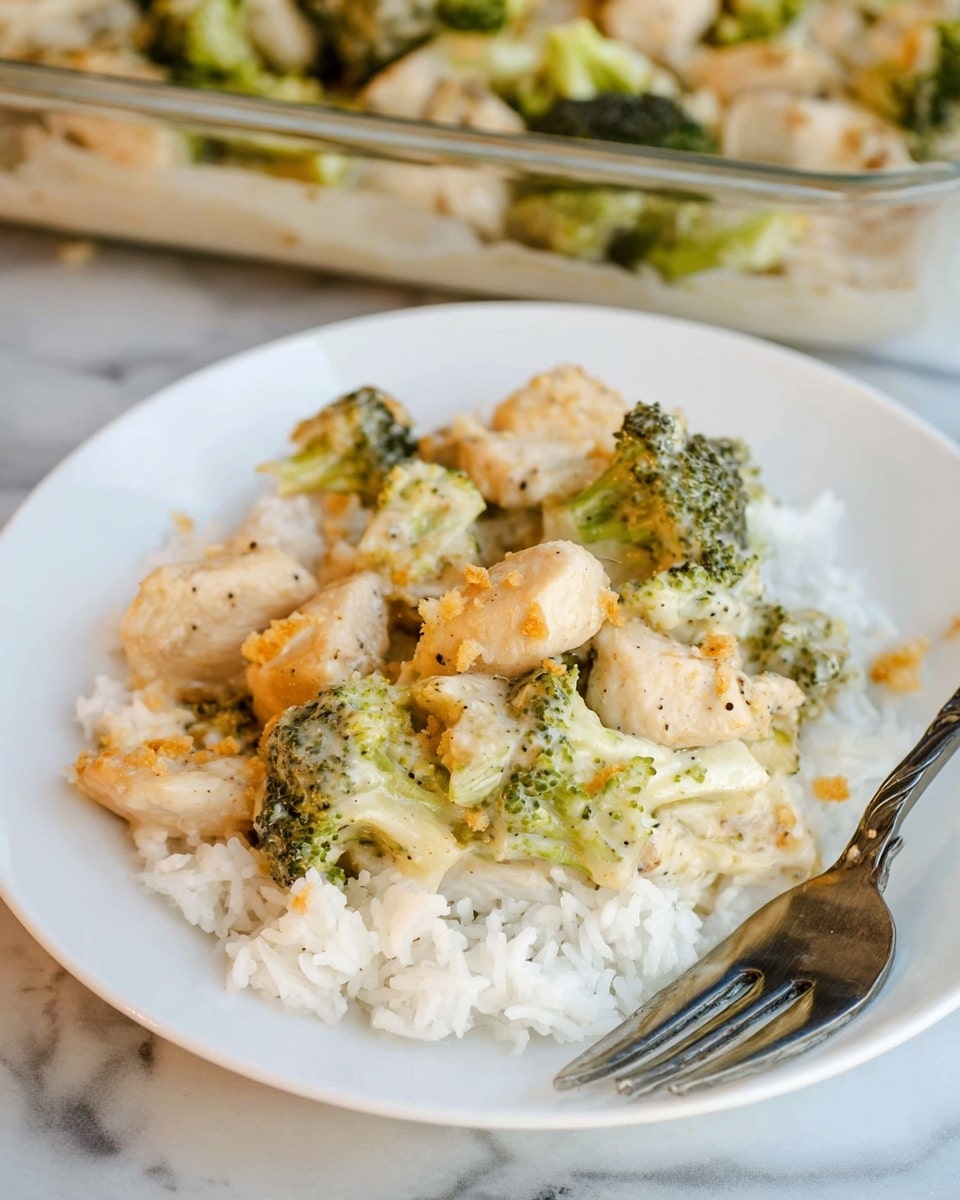 A white plate holds a serving of white rice as the bottom layer, topped with a creamy chicken and broccoli mixture as the second layer. The chicken pieces are light golden brown with a soft texture, and the broccoli florets are vibrant green with a slightly soft texture from the sauce. The creamy sauce is light beige with black pepper specks and some golden crumbs on top of the chicken. A silver fork rests on the right side of the plate. The plate sits on a white marbled surface with a glass baking dish of the same chicken and broccoli mix blurred in the background. Photo taken with an iphone --ar 4:5 --v 7