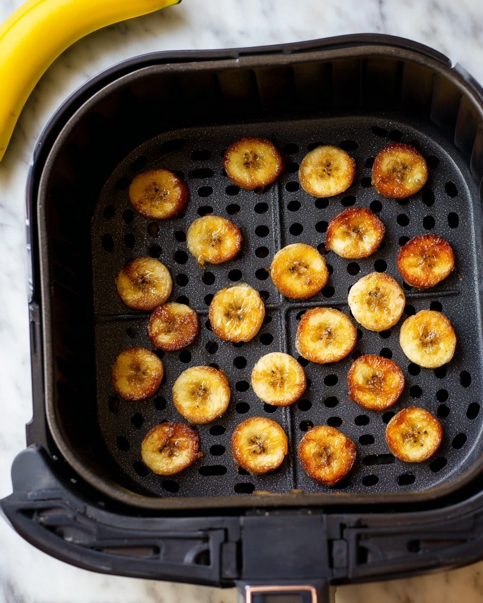 The image shows a dark round perforated tray inside an air fryer basket with about twenty small plantain slices. The plantain slices are cooked to a golden brown color with some darker caramelized spots, giving them a slightly crispy texture on top. The tray has small round holes allowing air flow and shows some cooking residue. In the upper part of the picture, two whole yellow bananas rest on a wooden cutting board. The overall setup is on a white marbled surface. photo taken with an iphone --ar 4:5 --v 7