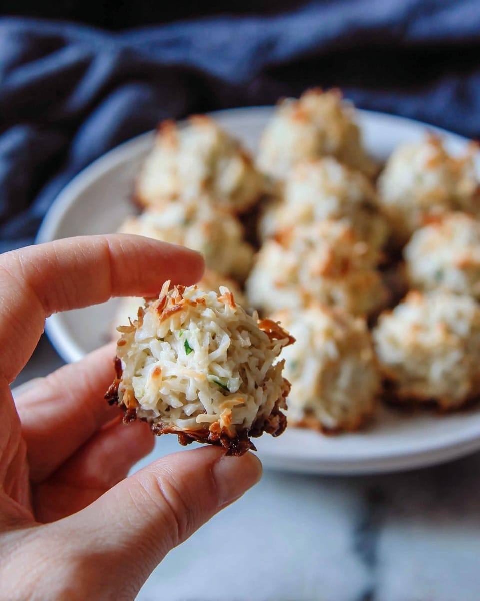 A close-up of a small round coconut macaroon with a rough, textured surface made of shredded white coconut, showing a golden brown crust on top and at the edges with a soft, moist white inside. The macaroon is held by a woman's hand in the front, and in the background, many similar macaroons are arranged closely together on a large white plate. The scene is set against a white marbled texture surface with a dark cloth in the background, creating contrast with the light-colored treats. photo taken with an iphone --ar 4:5 --v 7