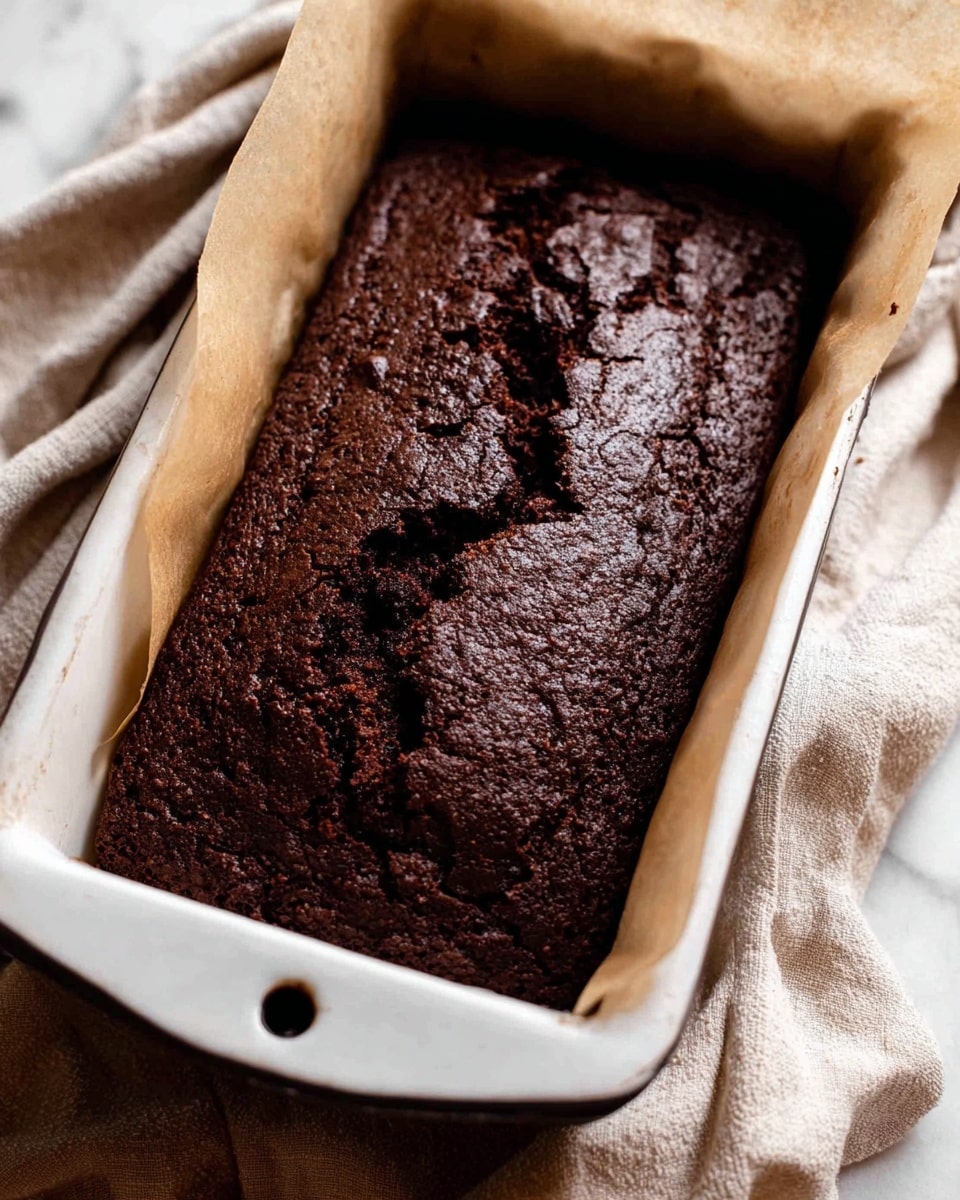 A close-up view of a dark brown chocolate loaf cake with a cracked, textured top surface sitting inside a white baking pan with a blue rim. The pan is lined with white parchment paper that extends over the sides. The background shows a soft beige fabric and a white marbled surface. The cake looks moist and rich with uneven cracks running across the top. photo taken with an iphone --ar 4:5 --v 7